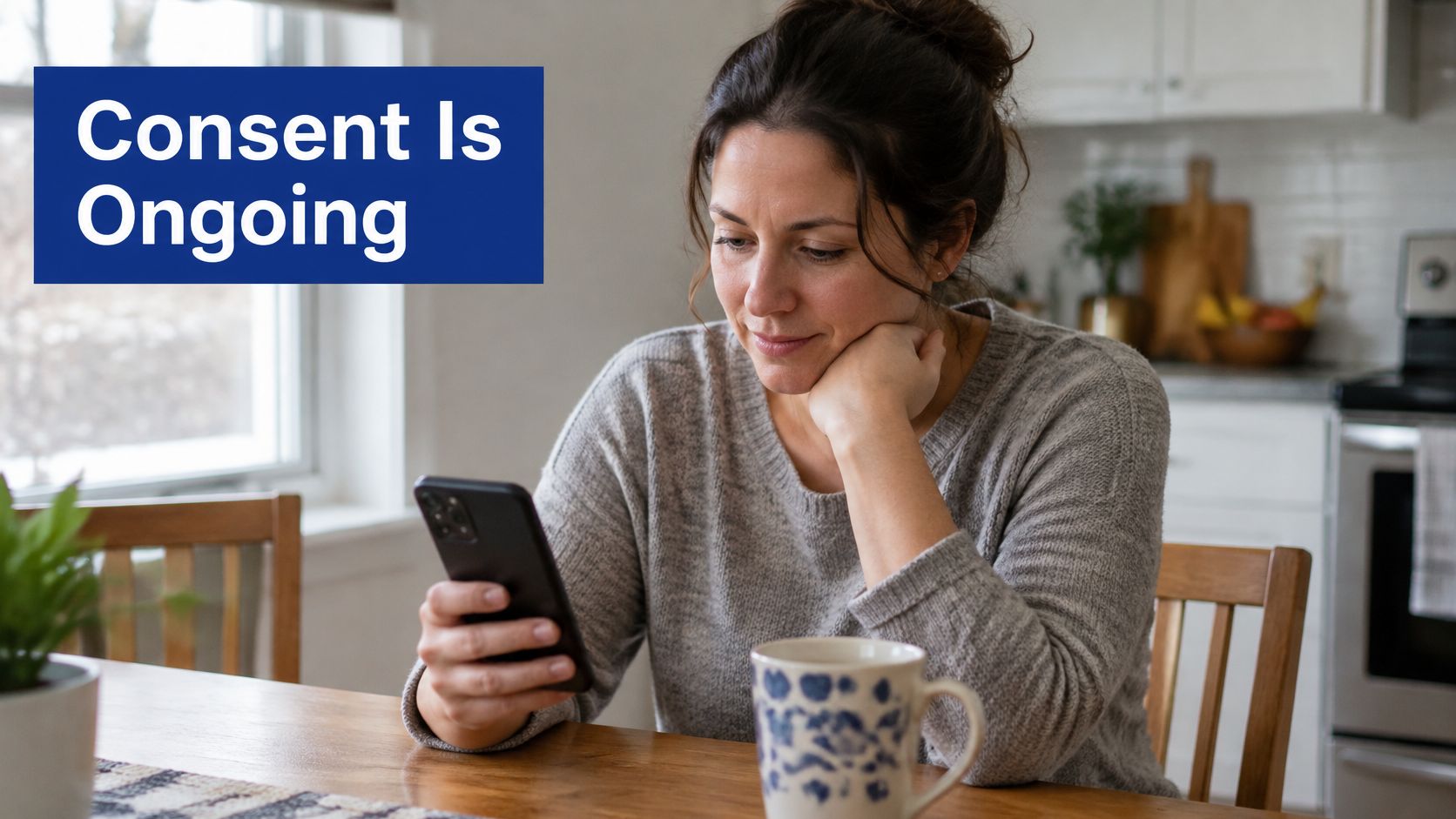 A thoughtful woman sits at a kitchen table using her smartphone with the words Consent Is Ongoing overlayed.